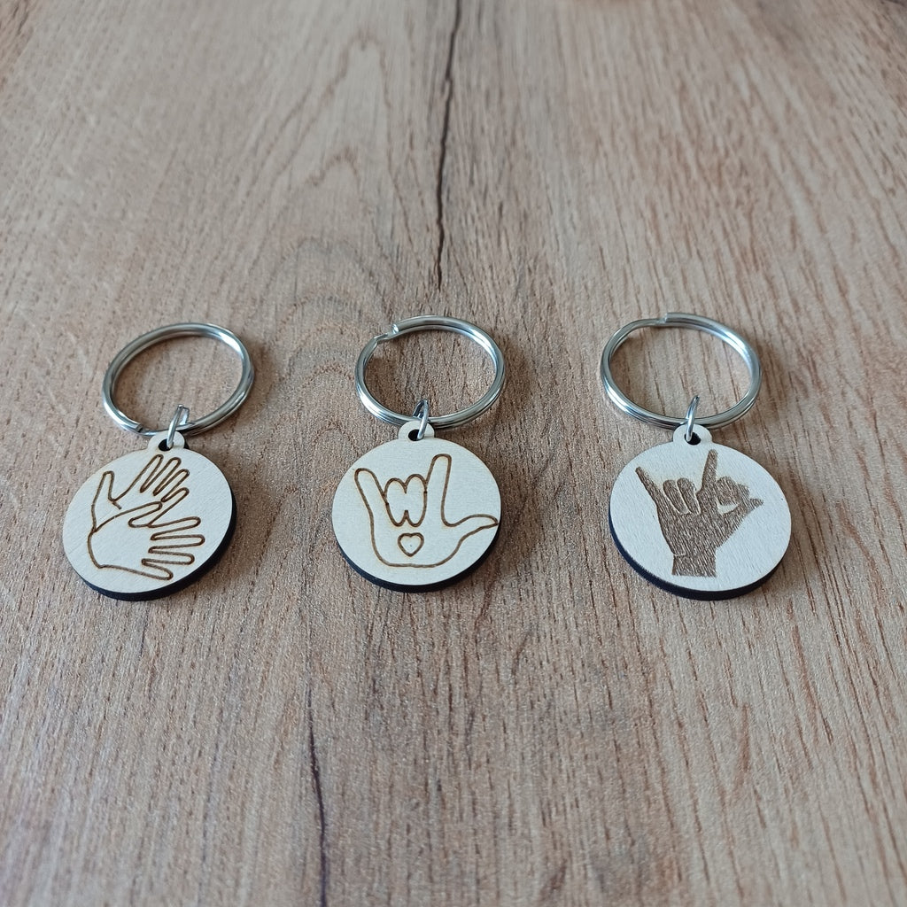 Trois mini porte-clés ronds en bois gravés en Langue des Signes LSF posés sur une table