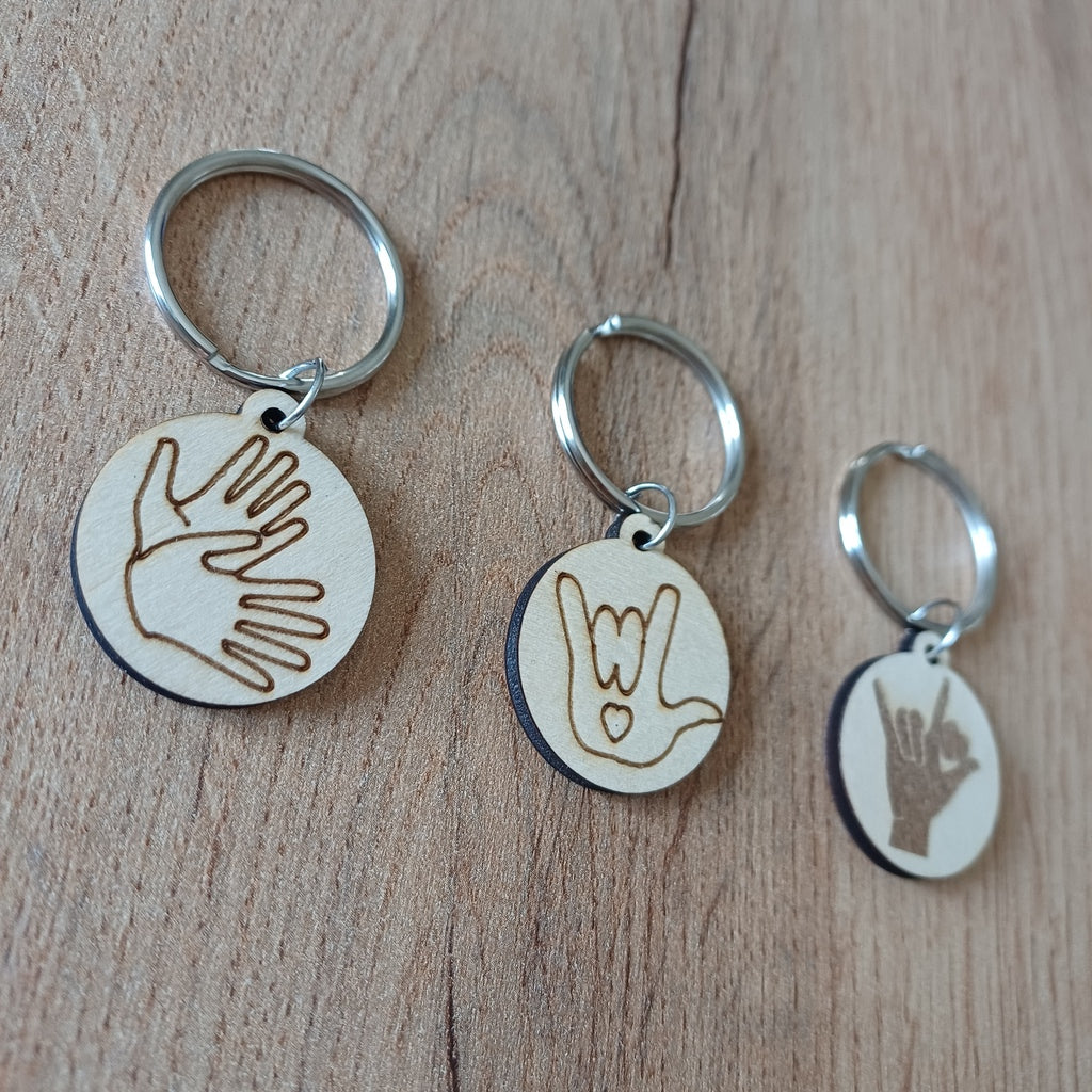 Trois mini porte-clés ronds en bois gravés en Langue des Signes LSF posés sur une table