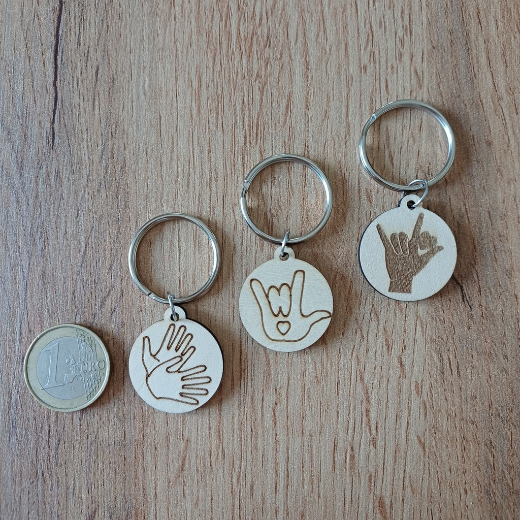 Trois mini porte-clés ronds en bois gravés en Langue des Signes LSF posés sur une table à côté d'une pièce de 1 euro pour montrer leur petite taille